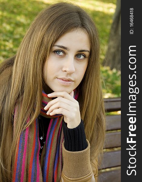 Vertical portrait of a beautiful young woman sitting on a bench in the park around a lot of yellow, red green leaves. Rests his head on his hand. Vertical portrait of a beautiful young woman sitting on a bench in the park around a lot of yellow, red green leaves. Rests his head on his hand