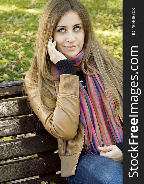 Young Caucasian woman calls on a mobile phone, sitting in a park on a wooden bench. Autumn around a lot of colorful foliage. Young Caucasian woman calls on a mobile phone, sitting in a park on a wooden bench. Autumn around a lot of colorful foliage