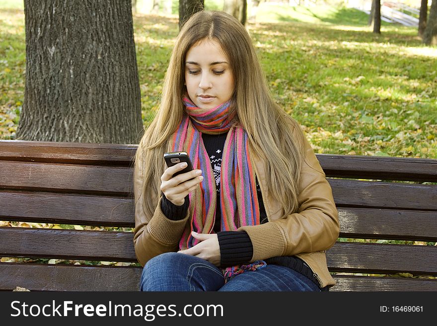 Young Caucasian woman with a cell phone, sitting in a park on a wooden bench, reading a SMS. Autumn around a lot of colorful foliage. Young Caucasian woman with a cell phone, sitting in a park on a wooden bench, reading a SMS. Autumn around a lot of colorful foliage