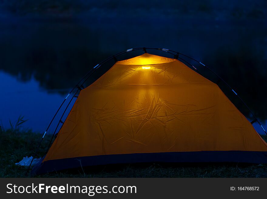 An orange tent at night stands on the shore and glows inside.