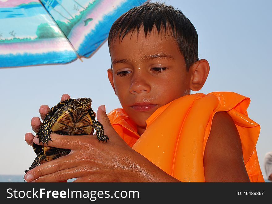 Boy with a turtle in his hands, wearing a vest to swim at the beach, on the background of sea and sun umbrella