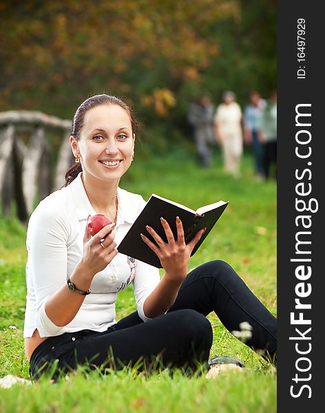 Youn smiling woman sitting on green grass with book and apple