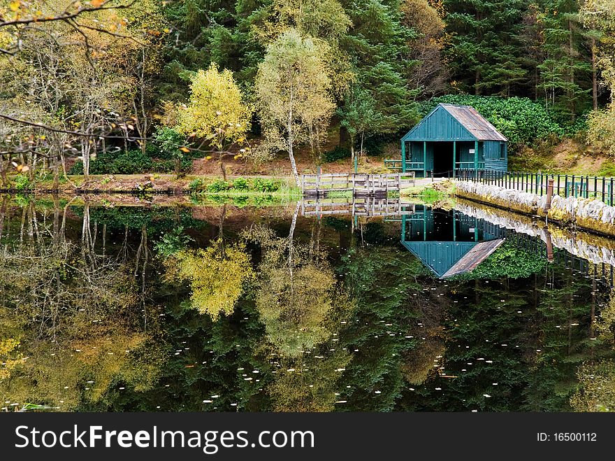 A horizontal image of a small loch in Scotland surrounded by trees and mountains with high reflective qualities in the water. A horizontal image of a small loch in Scotland surrounded by trees and mountains with high reflective qualities in the water