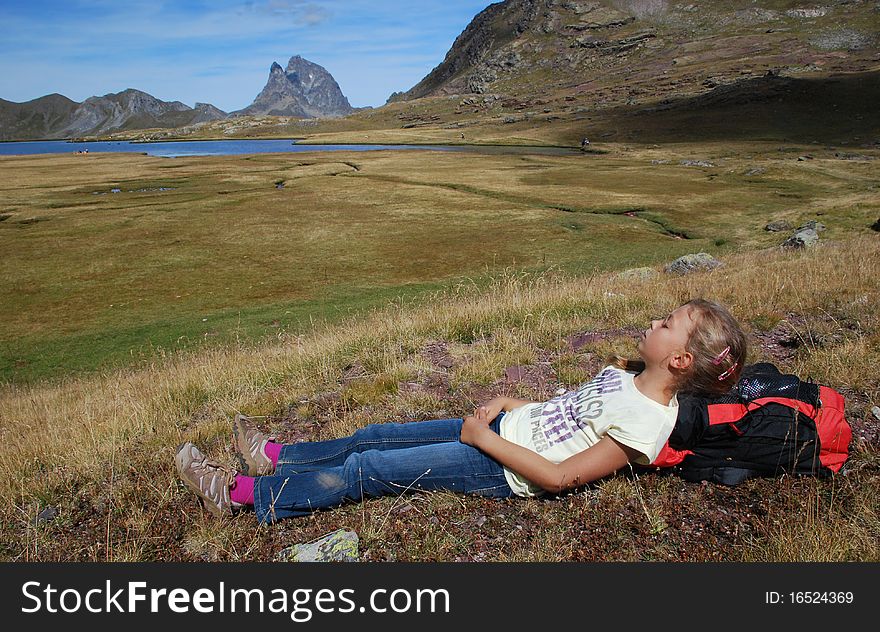 Small girl in focus, repose after walk, had on backpack, at Anayet plateau in Aragon, Spain. the lake and Beautiful peak du Midi d'Ossau in French site of Pytenees is at background. Small girl in focus, repose after walk, had on backpack, at Anayet plateau in Aragon, Spain. the lake and Beautiful peak du Midi d'Ossau in French site of Pytenees is at background