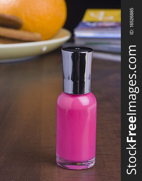 Pink nail polish standing on a table in front of a stack of magazines and a plate with food.