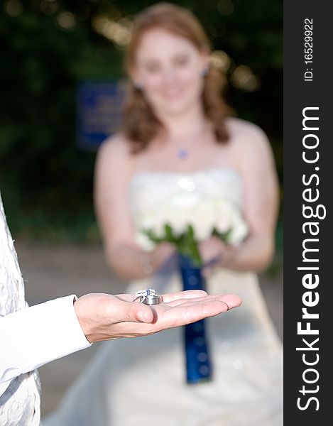 Wedding rings on the grooms palm with the bride smiling and holding flowers in the background