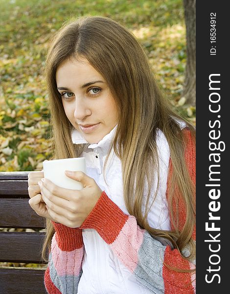 Beautiful young woman in autumn park sitting on a bench in the hands of white cup with coffee / tea. Beautiful young woman in autumn park sitting on a bench in the hands of white cup with coffee / tea