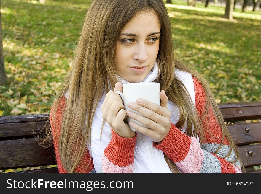 Beautiful young woman in autumn park sitting on a bench in the hands of white cup with coffee / tea. Beautiful young woman in autumn park sitting on a bench in the hands of white cup with coffee / tea