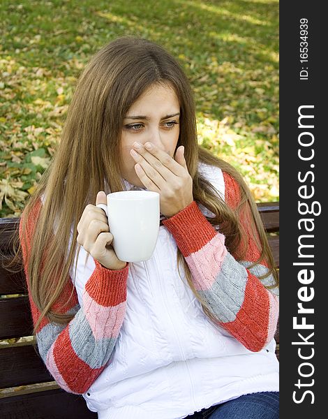 Beautiful young woman in autumn park sitting on a bench in the hands of white cup with coffee / tea. Beautiful young woman in autumn park sitting on a bench in the hands of white cup with coffee / tea