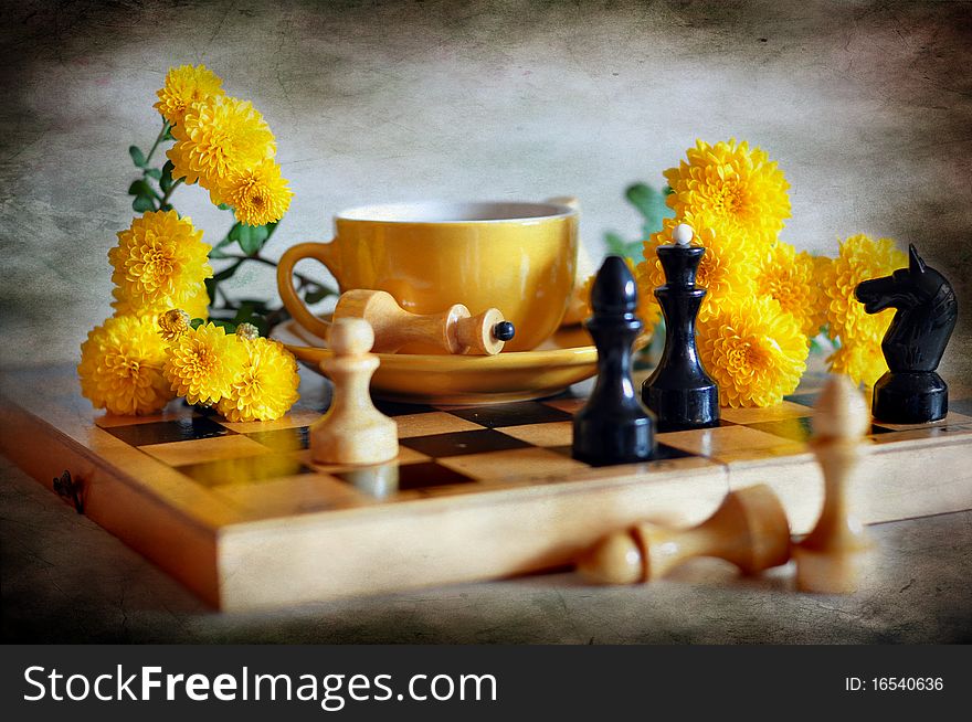 a cup with tea stands on a chess-board, alongside yellow chrysanthemums and chess figures. a cup with tea stands on a chess-board, alongside yellow chrysanthemums and chess figures