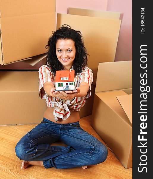 Attractive young woman offering a miniature house, boxes in the background. Attractive young woman offering a miniature house, boxes in the background