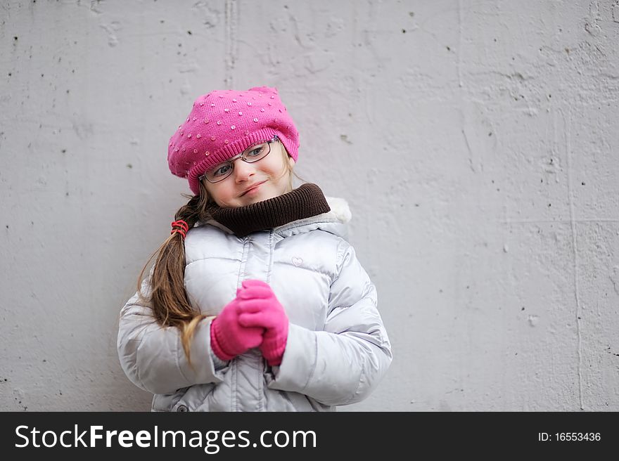 Adorable small girl in bright pink hat with long dark hair and glasses on grey background