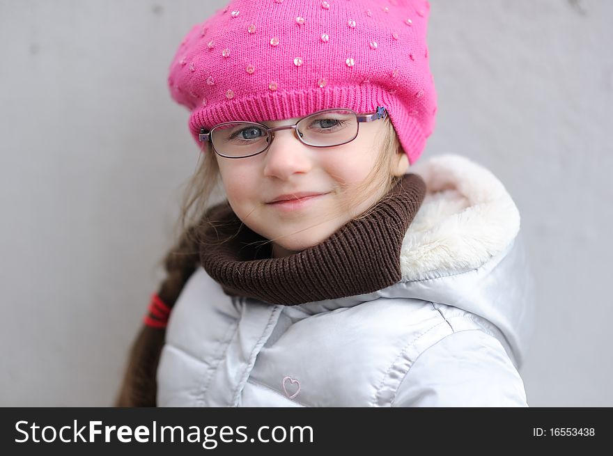 Adorable small girl in bright pink hat with long dark hair and glasses on grey background