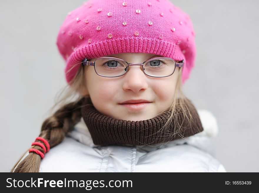 Adorable small girl in bright pink hat with long dark hair and glasses on grey background