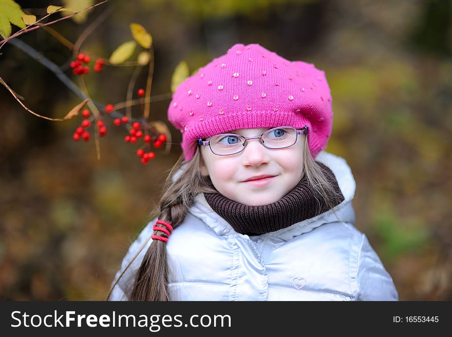 Adorable small girl in bright pink hat with long dark hair and glasses on beauty autumn background with rowanberry on it