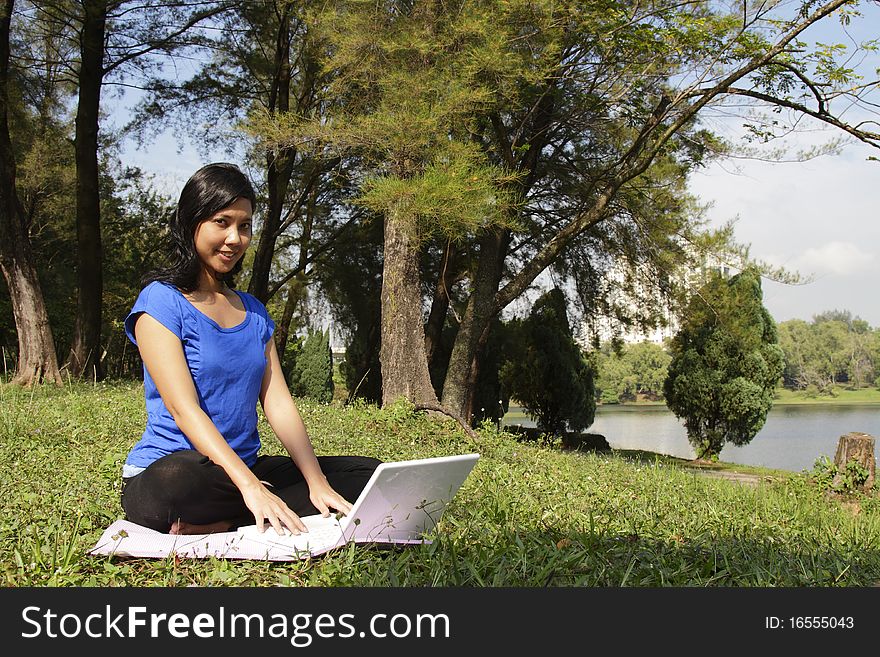 A smiling Asian woman with a laptop sitting on the ground at a park. A smiling Asian woman with a laptop sitting on the ground at a park