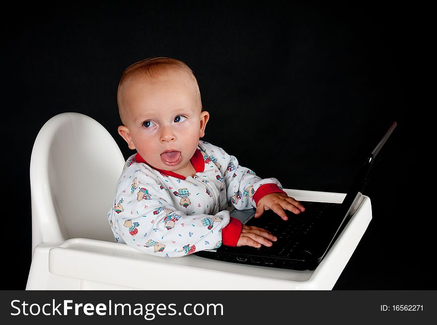 Baby sitting in the high chair, sticking out his tongue and using the laptop. Baby sitting in the high chair, sticking out his tongue and using the laptop