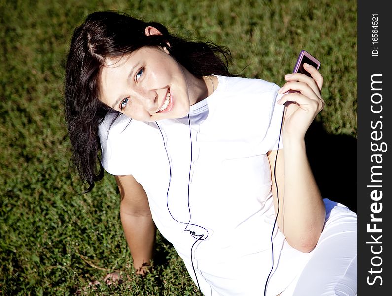 Pretty young brunette girl listening music on green grass in the park and show pink music player. Pretty young brunette girl listening music on green grass in the park and show pink music player.