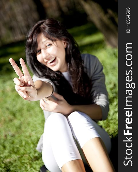 Portrait of beautiful brunette girl with blue eyes on green grass in the park show V symbol. Portrait of beautiful brunette girl with blue eyes on green grass in the park show V symbol.