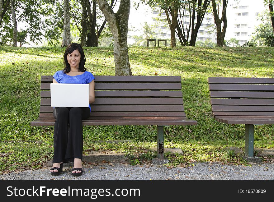 An Asian woman with a laptop sitting on a bench at a park. An Asian woman with a laptop sitting on a bench at a park