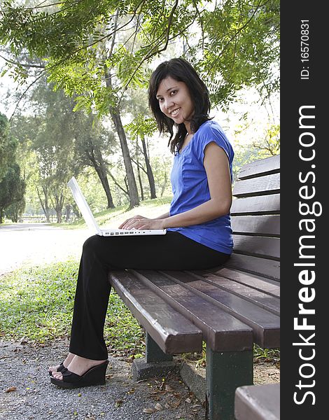 An Asian woman with a laptop sitting on a bench at a park. An Asian woman with a laptop sitting on a bench at a park