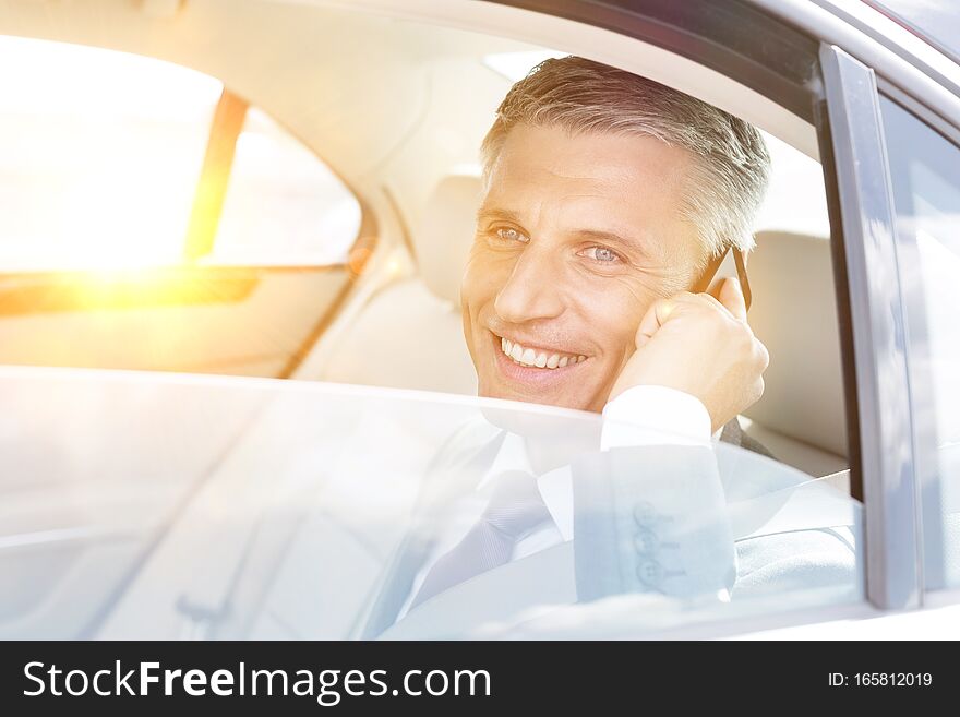 Photo of Smiling mature businessman talking on smartphone while looking through window in car with yellow lens flare in background