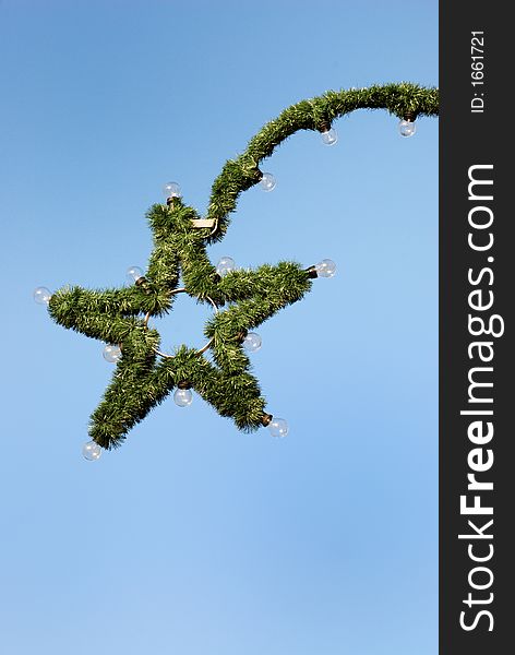 The picture shows a christmas star made of green fir branches. Attached are light bulbs.  In the background is blue sky. Image with copy space.