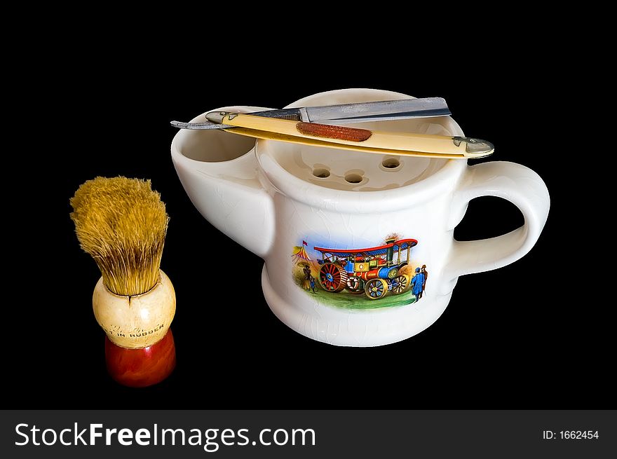 Close up of a white antique shaving mug with a handle on one end, a brush holder on the other, a 1890s scene on the side, a brush and a straight razor. Close up of a white antique shaving mug with a handle on one end, a brush holder on the other, a 1890s scene on the side, a brush and a straight razor.