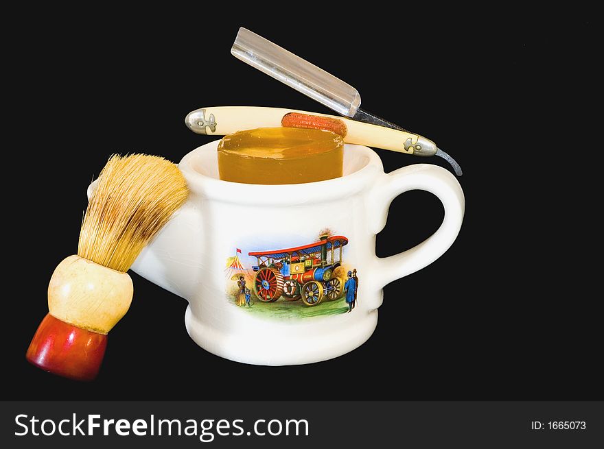 A white antique shaving mug with a handle on one end, a brush holder on the other, a 1890s scene on the side, a brush and a straight razor, all on a black background. A white antique shaving mug with a handle on one end, a brush holder on the other, a 1890s scene on the side, a brush and a straight razor, all on a black background.