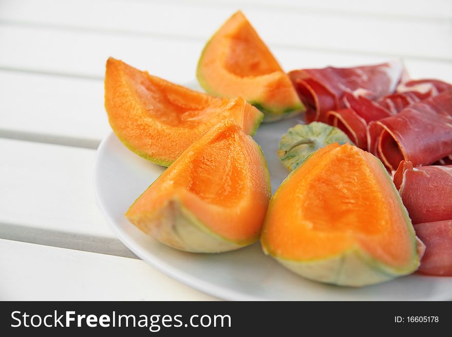 Slices of melon with italian ham in plain white plate on white picnic table. Slices of melon with italian ham in plain white plate on white picnic table.