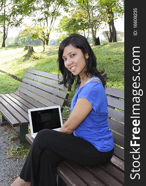 An Asian woman using a laptop on a bench at a park. An Asian woman using a laptop on a bench at a park