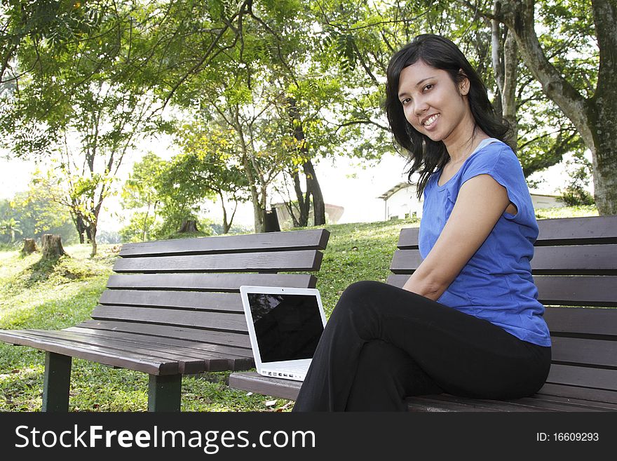 An Asian woman using a laptop on a bench at a park. An Asian woman using a laptop on a bench at a park