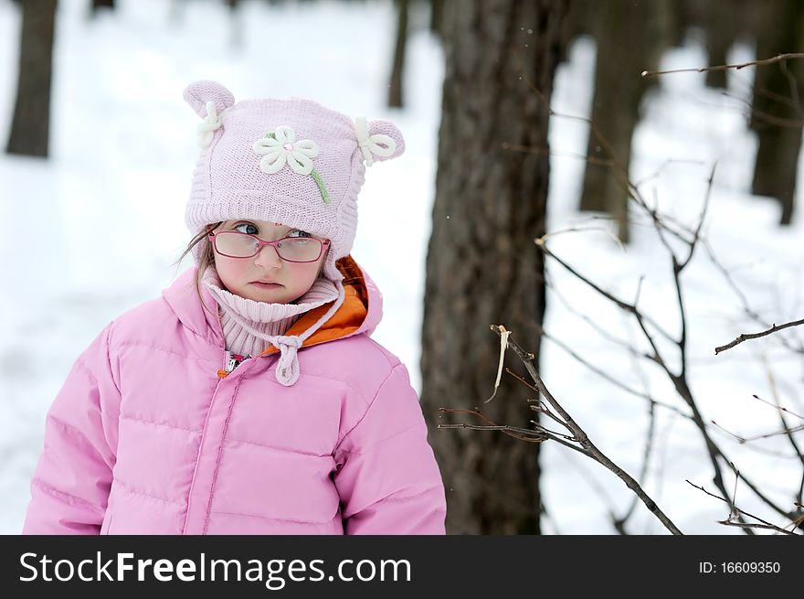 Adorable winter small girl in glasses and pink hat and jacket in the forest