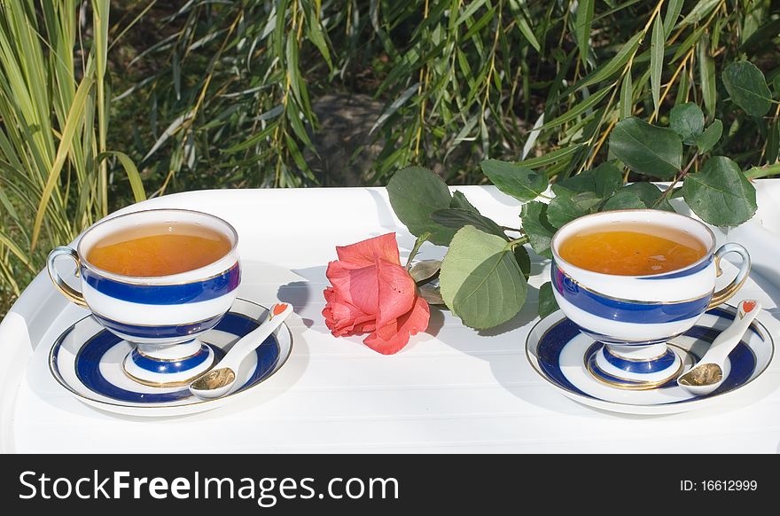 Two porcelain cups with tea on a white little table and a red rose in a summer garden against green trees