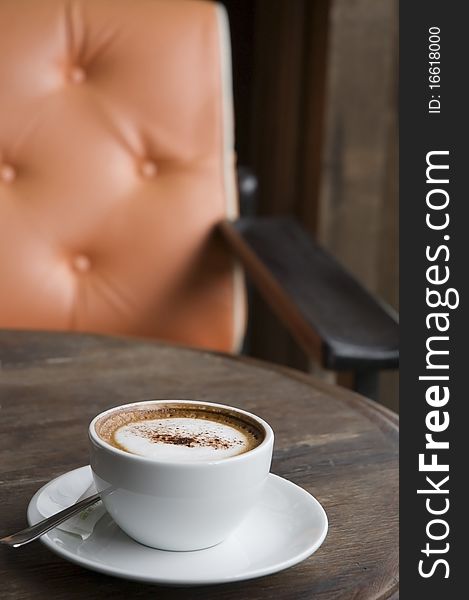 A cup of Cappuccino on wooden table with orange armchair background. A cup of Cappuccino on wooden table with orange armchair background
