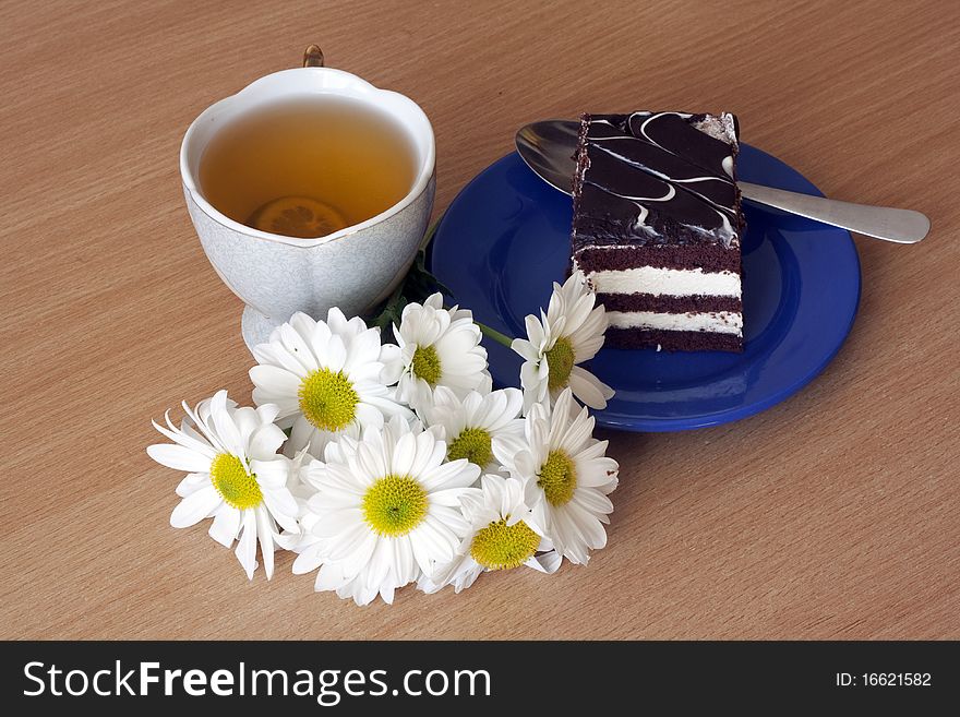 A cup of tea with lemon and a slice of chocolate cake on a plate next to a bouquet of chrysanthemums. A cup of tea with lemon and a slice of chocolate cake on a plate next to a bouquet of chrysanthemums