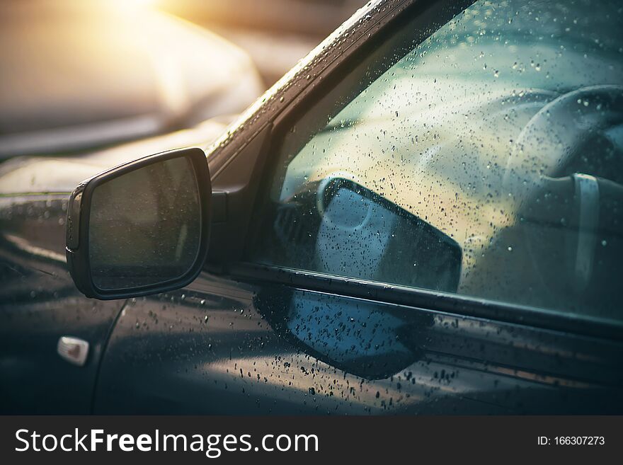 The rain-soaked windows and mirrors of the car, which are illuminated by the light of the sun that has just come out from behind the clouds