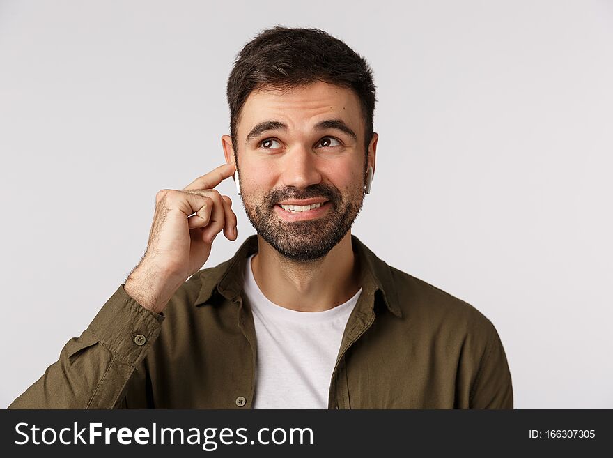 Carefree smiling handsome bearded adult male in coat, looking up dreamy and joyful, touching wireless earphone to tune