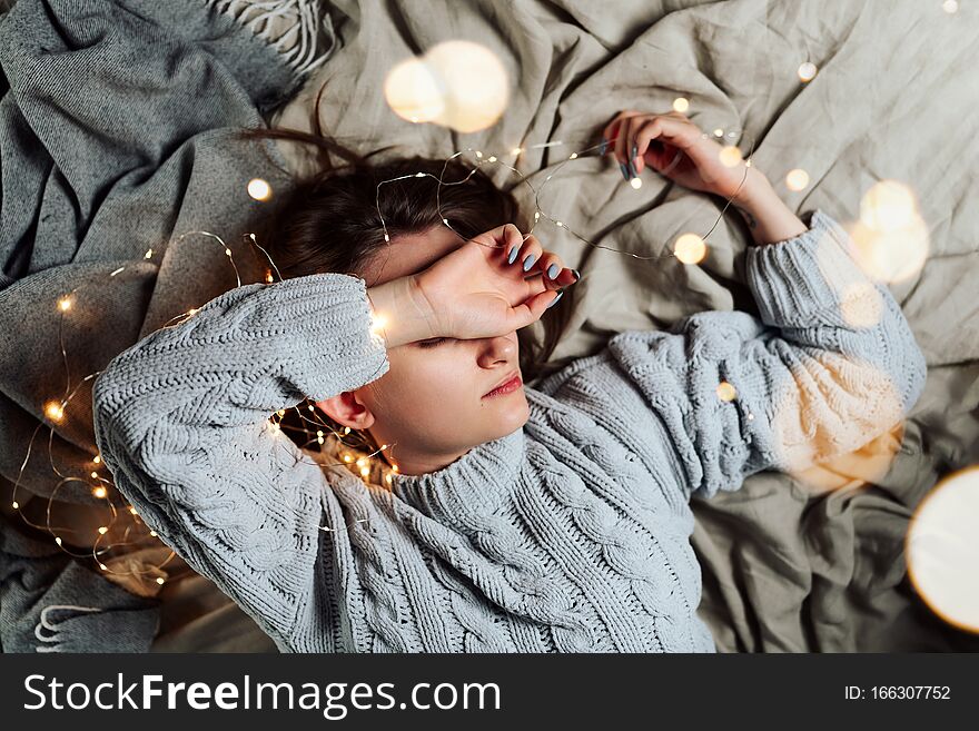 Young white woman lying in bed with fairy Christmas lights and bokeh around, shallow selective focus