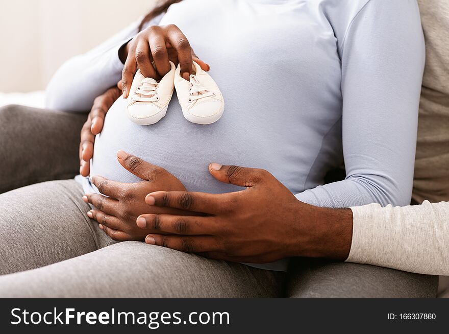 Close up of pregnant black woman belly with baby shoes on it, husband hugging his wife, new family concept. Close up of pregnant black woman belly with baby shoes on it, husband hugging his wife, new family concept