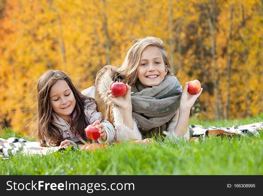 Beautiful caucasian big and little sisters lie on plaid, play with red apples on background of green grass of golden trees of autumn forest. Concept family weekend outdoors, people, lifestyle