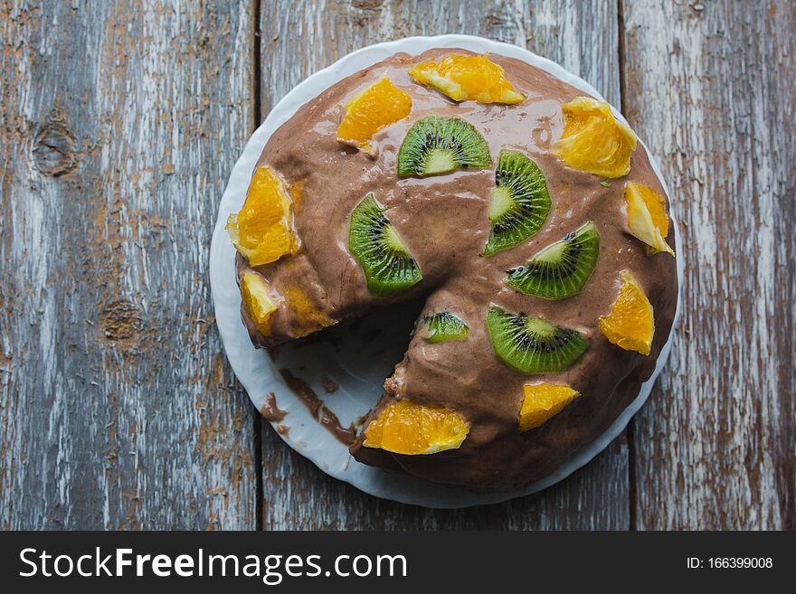 Homemade sponge cake with chocolate cacao ganache frosting. Slices of kiwi and orange on the dessert. Baked pastry in the kitchen on the old vintage wooden background.