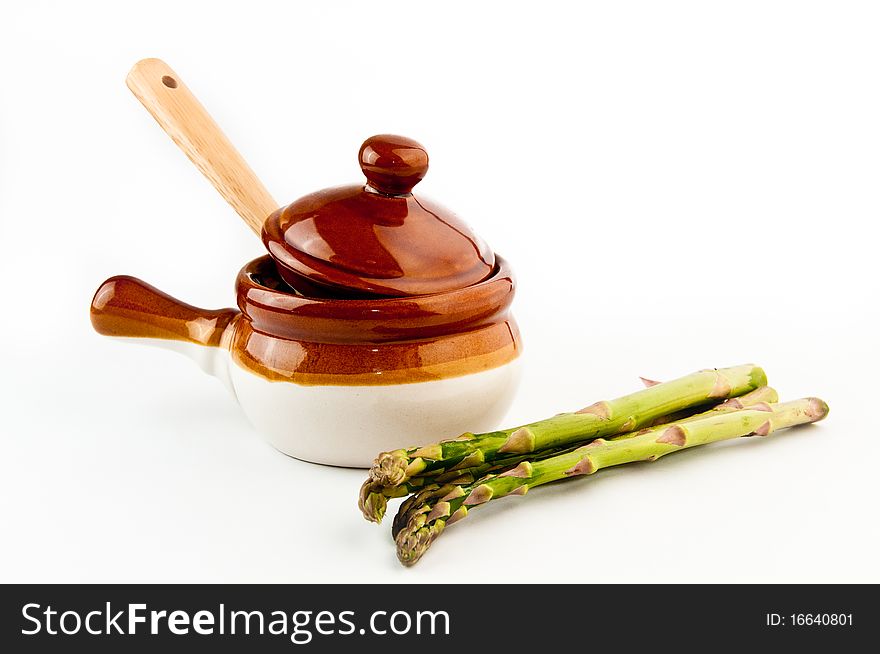 Brown soup crock with wooden spoon and fresh green asparagus on white background. Brown soup crock with wooden spoon and fresh green asparagus on white background