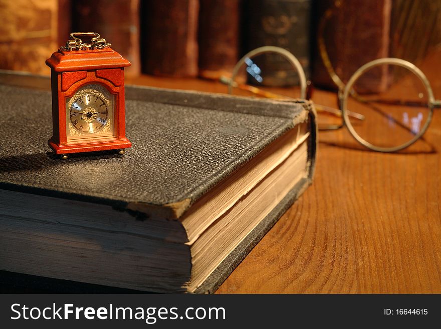 Vintage still life. Small wooden clock standing on old book on background with books and spectacles