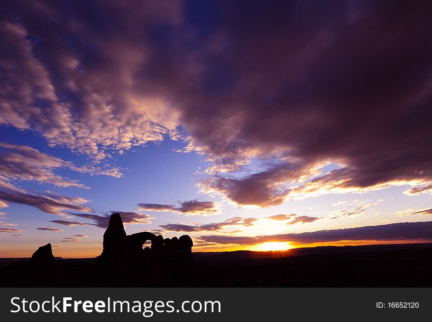 A dramatic sunset in Arches National Park casts a distant arch as a tranquil silhouette.