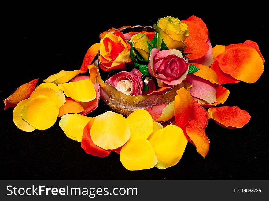 A silver basket with three artificial roses on orange, yellow and pink rose textile petals on black. A silver basket with three artificial roses on orange, yellow and pink rose textile petals on black