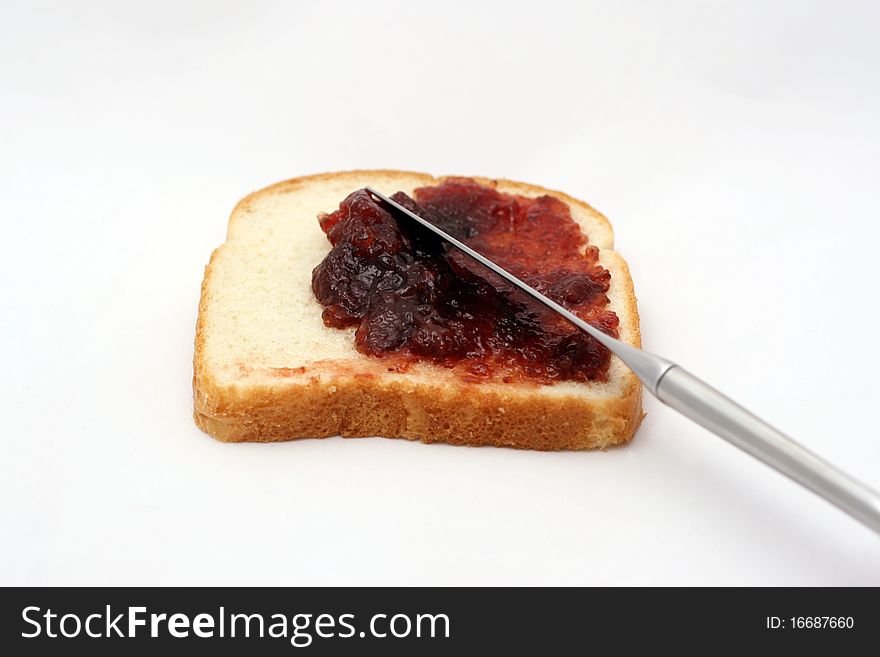 A piece of white bread with strawberry jelly being spread over it with a butter knife. A piece of white bread with strawberry jelly being spread over it with a butter knife.