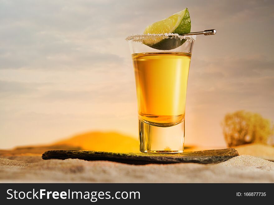 Gold tequila in a glass with salt and a slice of lime on a skewer stands on a stone against a beautiful sunset in the