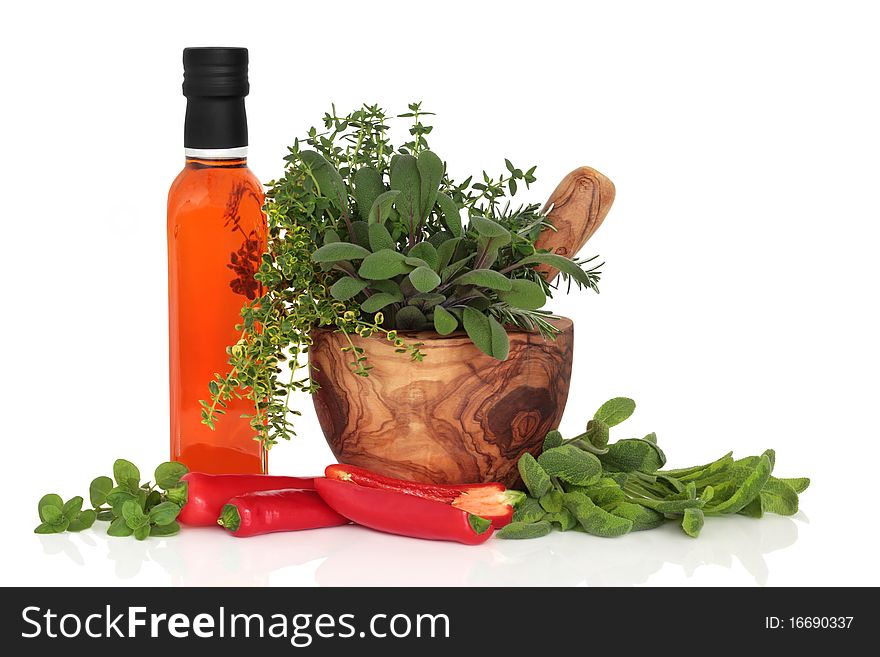 Chilli olive oil, chillies, and herb leaf sprigs of rosemary,  sage, thyme and oregano in an olive wood mortar with pestle, isolated over white background. Chilli olive oil, chillies, and herb leaf sprigs of rosemary,  sage, thyme and oregano in an olive wood mortar with pestle, isolated over white background.