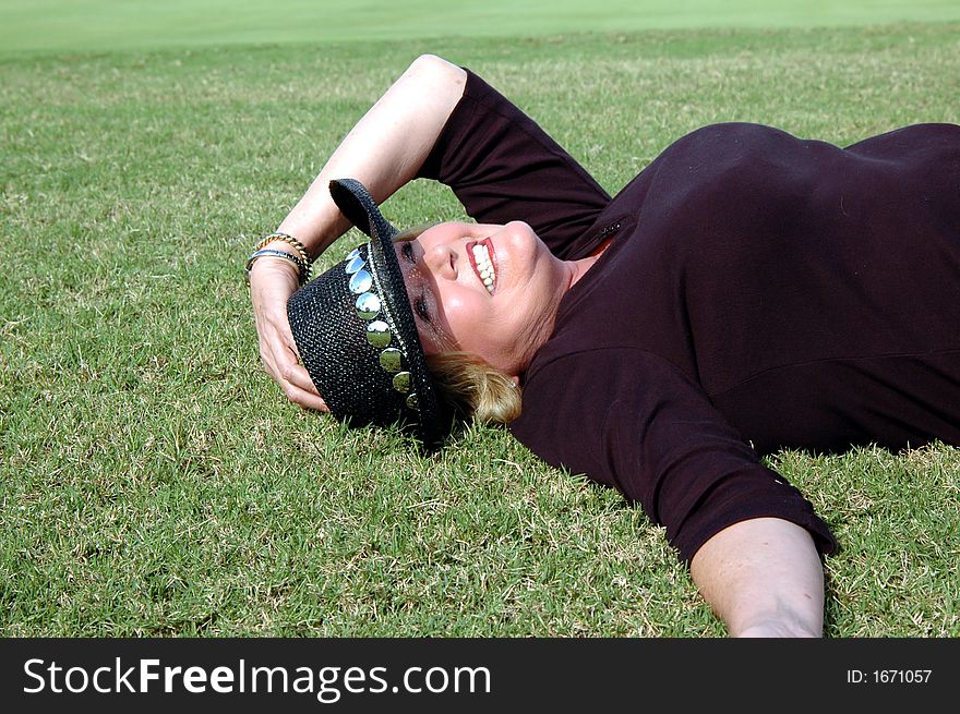 A smiling mature woman wearing a cowboy hat and lying down in the grass. Dressed in elbow length maroon sweater. Her left hand is holding her cowboy hat. . A smiling mature woman wearing a cowboy hat and lying down in the grass. Dressed in elbow length maroon sweater. Her left hand is holding her cowboy hat.